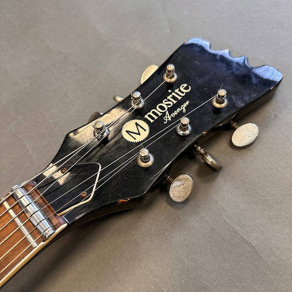 Close-up of black Mosrite Avenger guitar headstock with six tuning pegs and visible strings on gray background