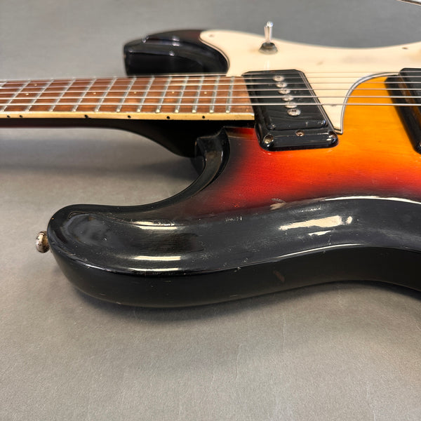Close-up of vintage electric guitar body with sunburst finish and cream pickguard, showing P-90 pickup and brown fretboard on gray background