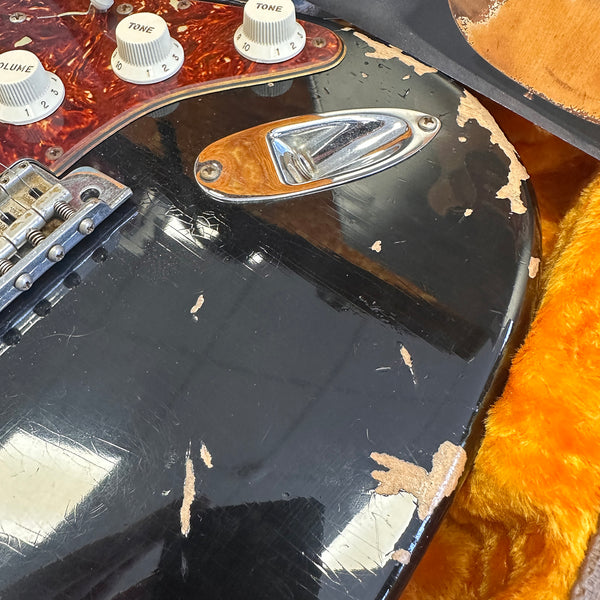 Close-up of vintage black electric guitar body showing worn finish, chrome input jack plate, tortoiseshell pickguard, tone and volume control knobs, and part of a metal bridge, resting on orange plush case lining.