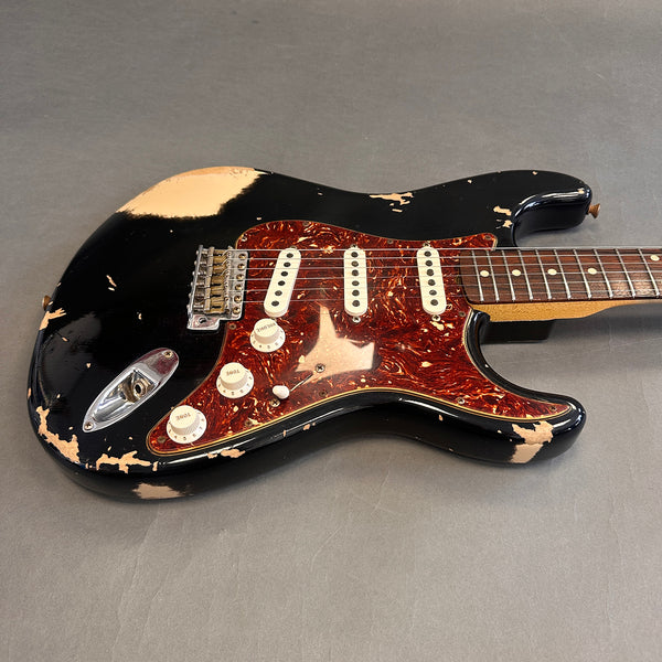Black electric guitar with heavy paint wear and tortoiseshell pickguard, featuring three single-coil pickups and white control knobs on gray background