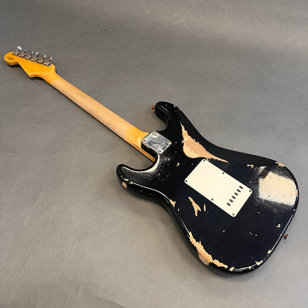 Black electric guitar with relic finish showing worn paint, viewed from the back, featuring a maple neck and chrome neck plate on gray background