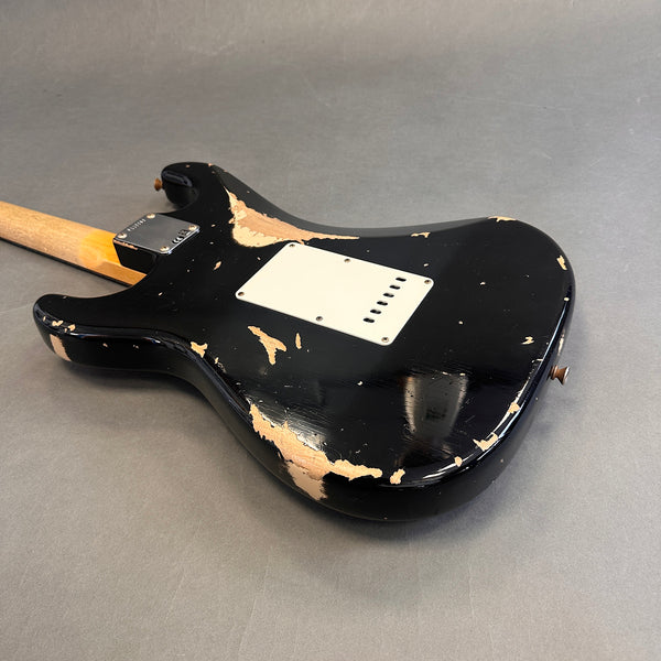 Black electric guitar body with worn, chipped paint revealing wood underneath, showing vintage relic wear and a white backplate on a gray surface.