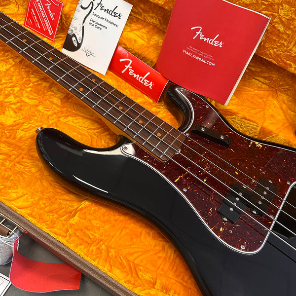Close-up of black Fender electric bass guitar with tortoiseshell pickguard in yellow plush-lined case, showing fretboard and included Fender manuals and warranty card.