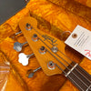 Close-up of Fender Precision Bass headstock with four tuning pegs and strings, resting in a yellow plush guitar case with a California Proposition 65 warning tag attached