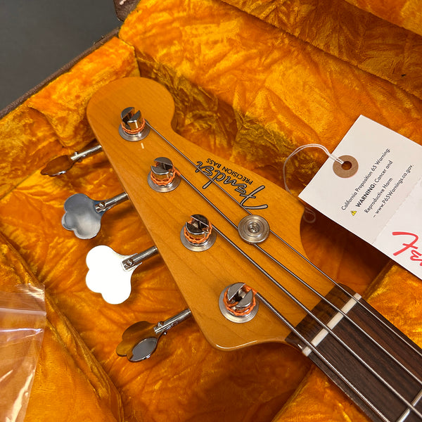 Close-up of Fender Precision Bass headstock with four tuning pegs and strings, resting in a yellow plush guitar case with a California Proposition 65 warning tag attached