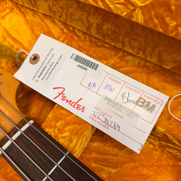Close-up of Fender guitar tag with assembly, tune/test, and inspector information, attached to a guitar neck with strings on an orange plush case interior background.