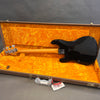 Black electric bass guitar with maple neck lying in open vintage-style orange plush-lined hard case on gray surface