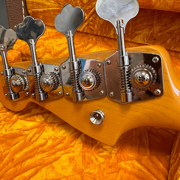 Close-up of tuning machines on the headstock of a natural wood electric bass guitar resting on orange plush fabric case lining
