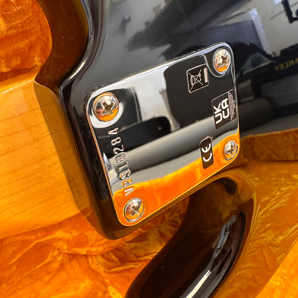 Close-up of guitar neck plate with serial number V218284, CE, and UKCA certification stamps on glossy black and natural wood finish guitar body.