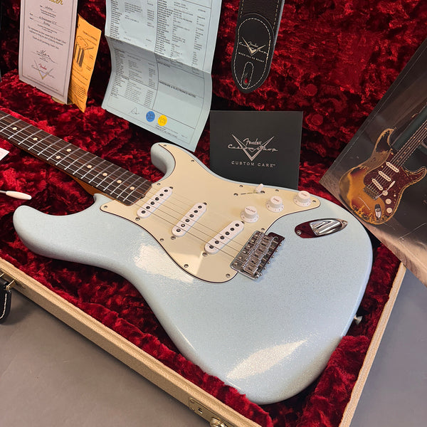 Fender electric guitar in light blue sparkle finish with white pickguard and three single-coil pickups, displayed in red plush-lined guitar case with Fender Custom Care documents and strap.