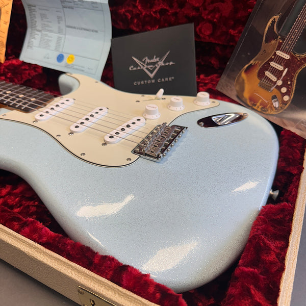 Fender Stratocaster electric guitar in light blue sparkle finish with white pickguard and three single-coil pickups, displayed in a red velvet-lined guitar case with Fender Custom Care booklet.