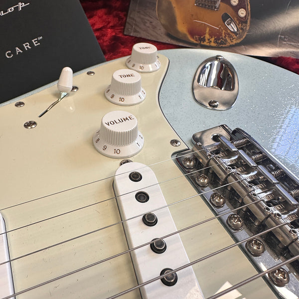 Close-up of white electric guitar body showing volume and tone control knobs, pickup, and chrome hardware components, with guitar care booklet and vintage guitar photo in background.