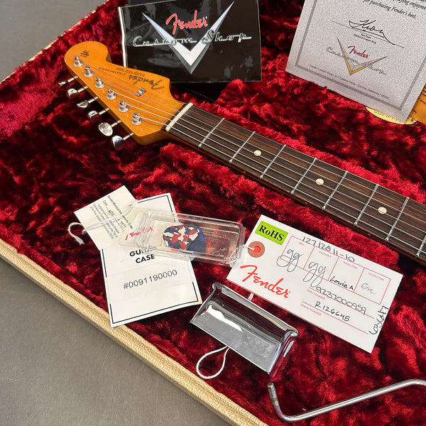 Fender Custom Shop guitar headstock and rosewood fretboard inside a red plush case with Fender warranty card, guitar pick in clear plastic case, metal tremolo cover, and Fender quality assurance tags.
