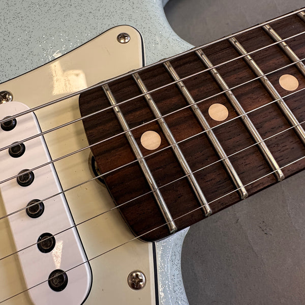 Close-up of electric guitar neck with rosewood fretboard, metal frets, and dot inlays next to white pickup and light blue speckled guitar body
