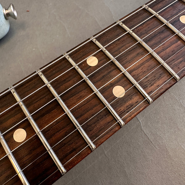 Close-up of guitar fretboard with metal frets and dot inlays on dark wood, guitar strings visible, shown on gray surface.