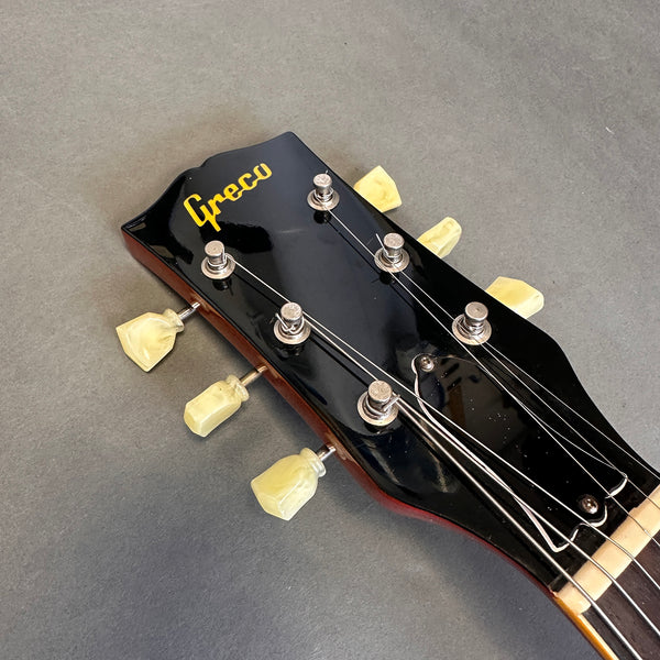 Close-up of black Greco guitar headstock with yellow tuning pegs and strings on gray background