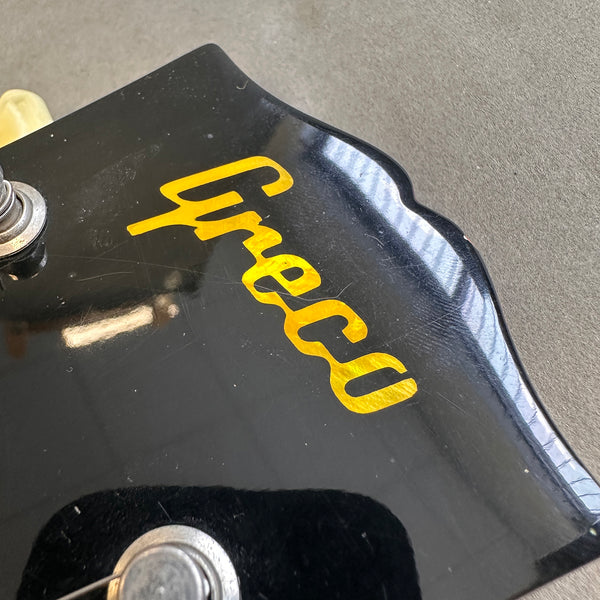 Close-up of Greco guitar headstock in black with yellow logo and tuning peg on gray background