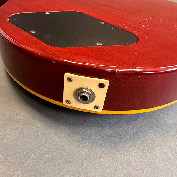 Close-up of red electric guitar body with yellow binding and black plastic control cavity cover, featuring output jack plate on gray surface.