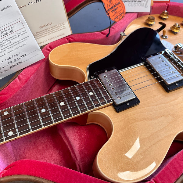Close-up of natural finish Gibson ES-335 electric guitar inside glossy red plush hard case with warranty and authenticity papers visible