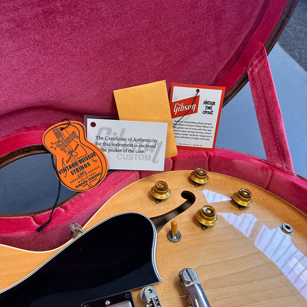 Close-up of semi-hollow electric guitar body with sunburst finish, showing black pickguard, gold control knobs, toggle switch, and partially visible f-hole, inside a plush pink-lined guitar case with Gibson vintage reissue strings tag and certificate of authenticity cards.
