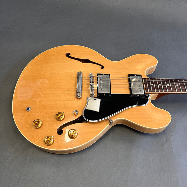 Semi-hollow electric guitar with natural wood finish, dual humbucker pickups, black pickguard, and four gold control knobs on gray background