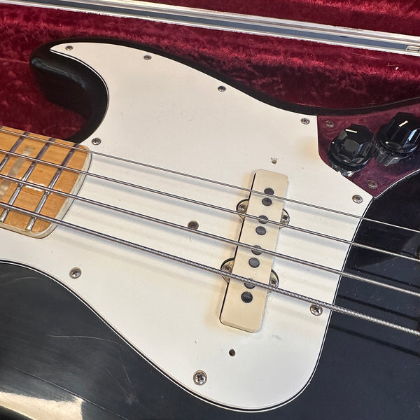 Close-up of black electric bass guitar body with white pickguard, chrome strings, maple fretboard, and black control knobs in a red velvet-lined case