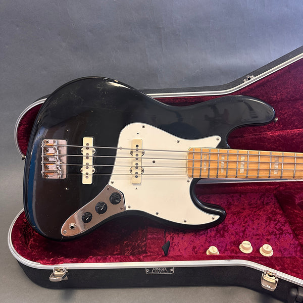 Black vintage electric bass guitar with white pickguard and maple neck resting in red velvet lined Hiscox hard case on gray background