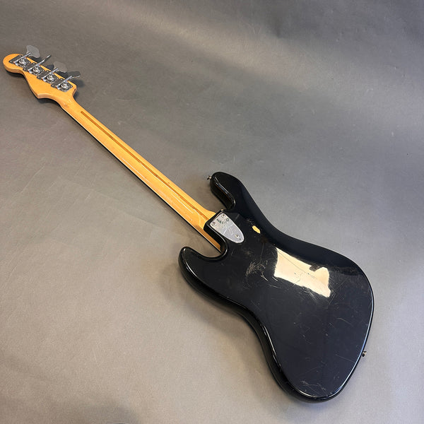 Black electric bass guitar with a maple neck and chrome tuning pegs, shown from the back on a gray surface, featuring visible wear and scratches on the body.