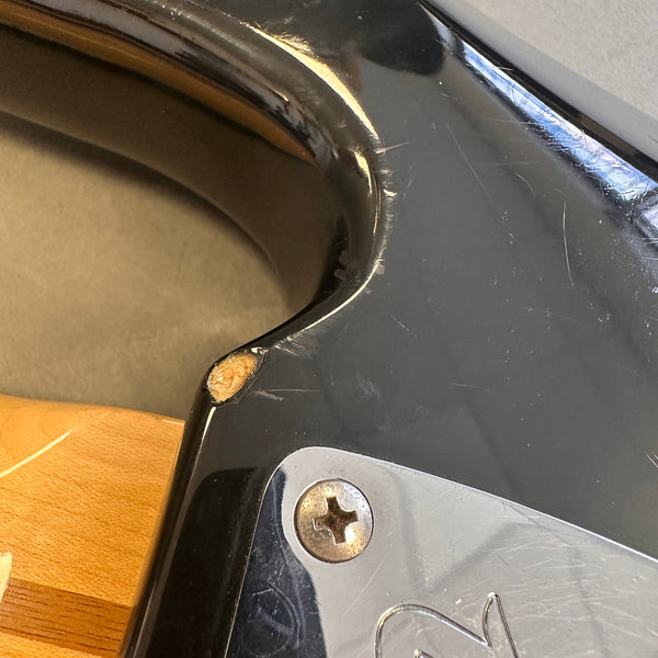 Close-up of black electric guitar body showing paint chip and scratches near neck joint and metal plate screw.