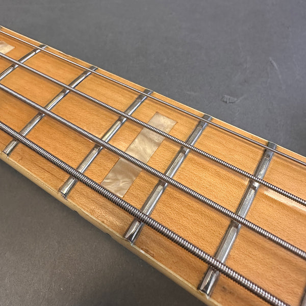 Close-up of maple bass guitar fretboard with metal frets, pearl block inlay, and round wound strings on grey background