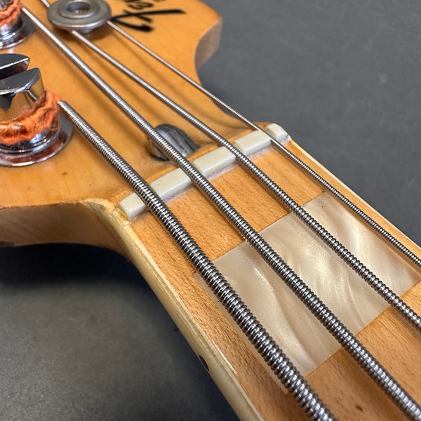 Close-up of bass guitar headstock showing tuning pegs, strings, and pearl block inlay on maple neck with black background
