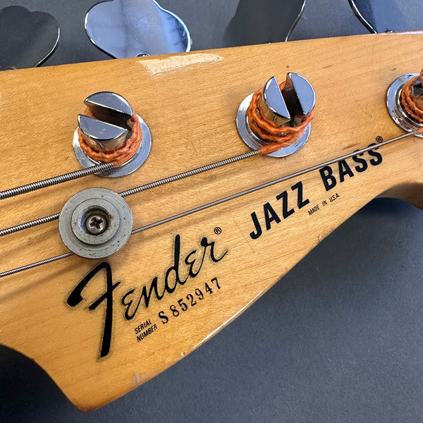 Close-up of Fender Jazz Bass headstock with serial number S852947 and tuning pegs on natural wood finish