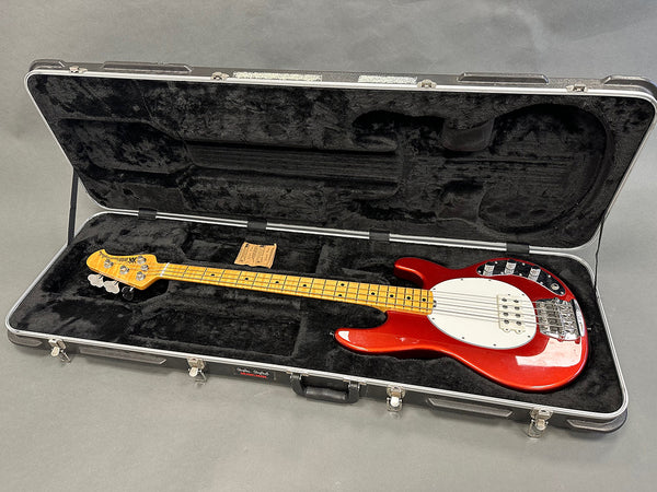Red electric bass guitar with maple neck and black hard-shell case interior, open case on grey background