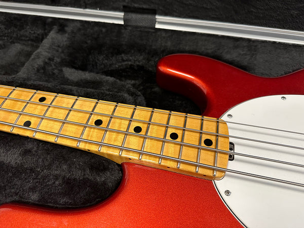 Close-up of maple fingerboard and red electric bass guitar body with white pickguard inside black plush case with bow accessory.