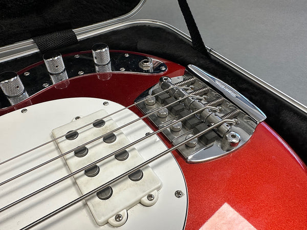 Close-up of red electric bass guitar body showing white pickup, chrome bridge, control knobs, and strings in hard case interior