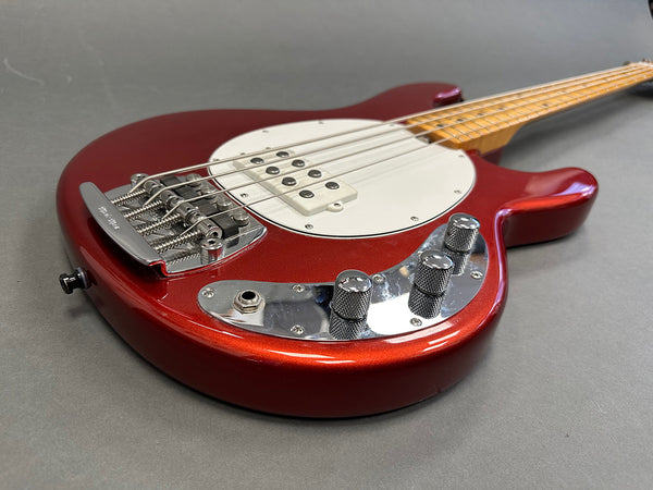 Close-up of red electric bass guitar body with white pickguard, chrome bridge, and control knobs on gray background
