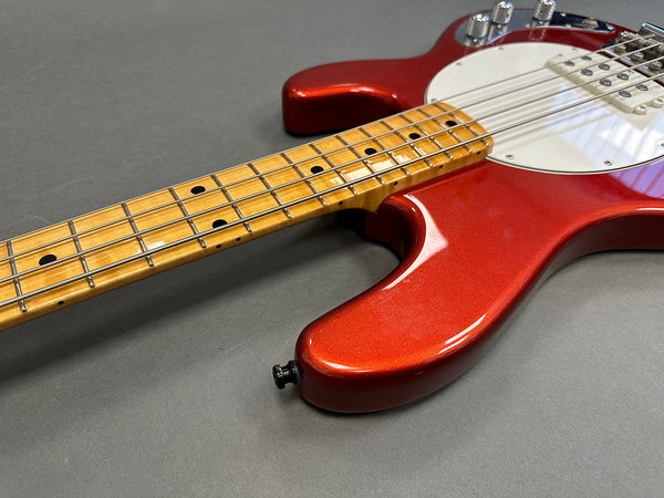 Close-up of electric bass guitar with metallic red body, maple neck, white pickguard, and chrome control knobs on gray background