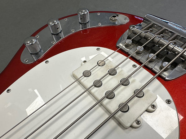 Close up of red electric bass guitar body showing white pickguard, chrome bridge, pickups, and control knobs