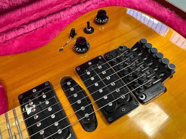 Close-up of honey burst electric guitar body showing three pickups, volume and tone knobs, selector switch, and Floyd Rose tremolo bridge with six strings over a pink plush guitar case interior.