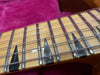 Close-up of vintage guitar fretboard with black trapezoid inlays, metal frets, and steel strings on natural wood neck background with purple fabric.