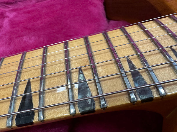 Close-up of vintage guitar fretboard with black trapezoid inlays, metal frets, and steel strings on natural wood neck background with purple fabric.