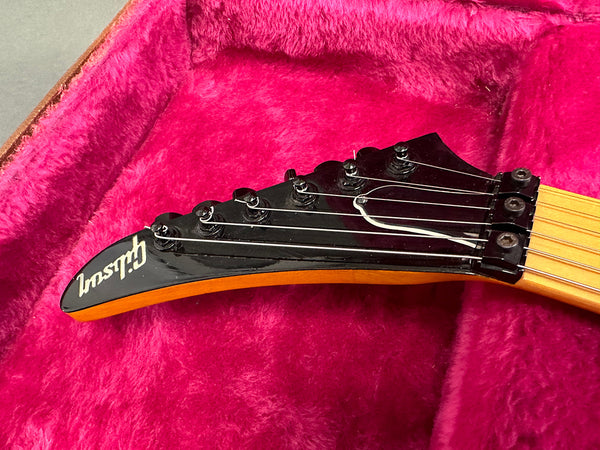 Close-up of black Gibson electric guitar headstock with six tuning pegs and strings, resting in a pink plush-lined guitar case.