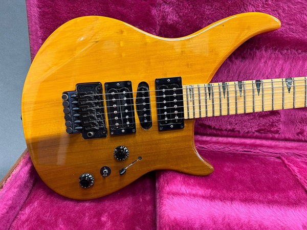 Electric guitar with natural wood finish, dual humbucker and single coil pickups, maple neck with sharktooth inlays, Floyd Rose locking tremolo in pink plush case
