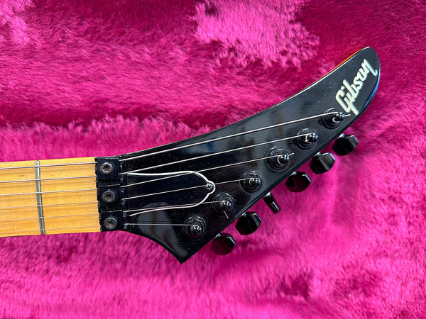Close-up of black Gibson electric guitar headstock with six tuning pegs and strings, on bright pink plush background