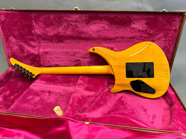 Back view of honey-colored electric guitar with black backplates inside pink plush-lined guitar case with guitar pick.