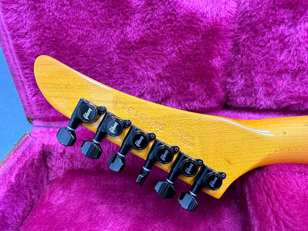 Close-up of Gibson guitar headstock with six black tuning pegs inside a purple plush-lined guitar case