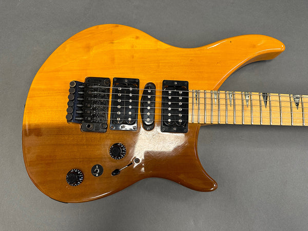 Electric guitar with honey amber finish, double humbucker pickups, single coil middle pickup, maple fretboard with shark tooth inlays, Floyd Rose tremolo system, and black control knobs on gray background.