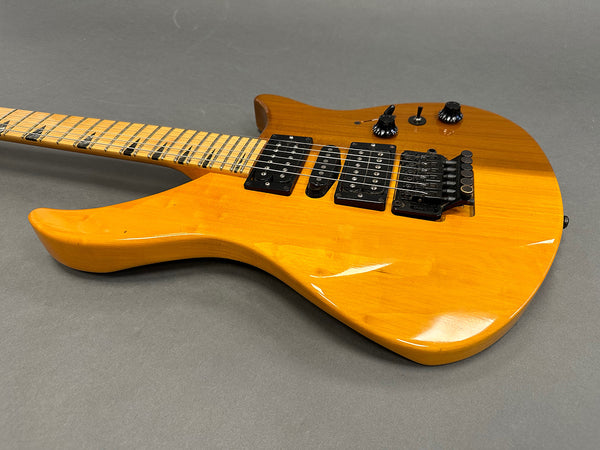 Electric guitar with natural wood finish, maple neck with black shark tooth inlays, dual humbucker and single coil pickups, and black hardware including a Floyd Rose tremolo system on gray background