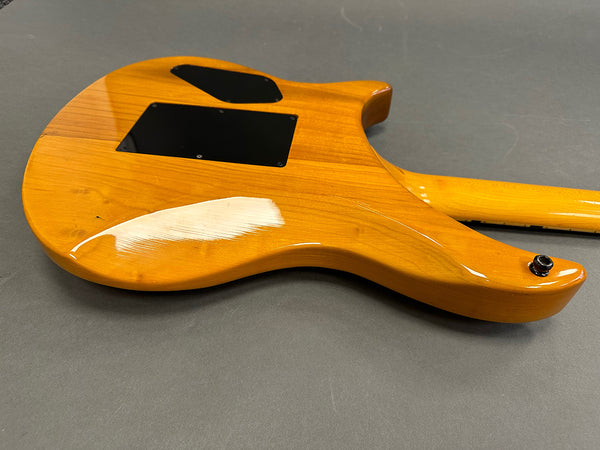 Back of natural wood electric guitar body with black plastic covers and strap button on gray surface