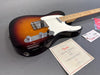 Fender electric guitar in sunburst finish with maple neck and chrome hardware alongside Fender certificate of authenticity and tags on gray background